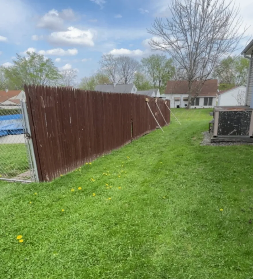 Old dark brown fence sagging across a green backyard with temporary braces